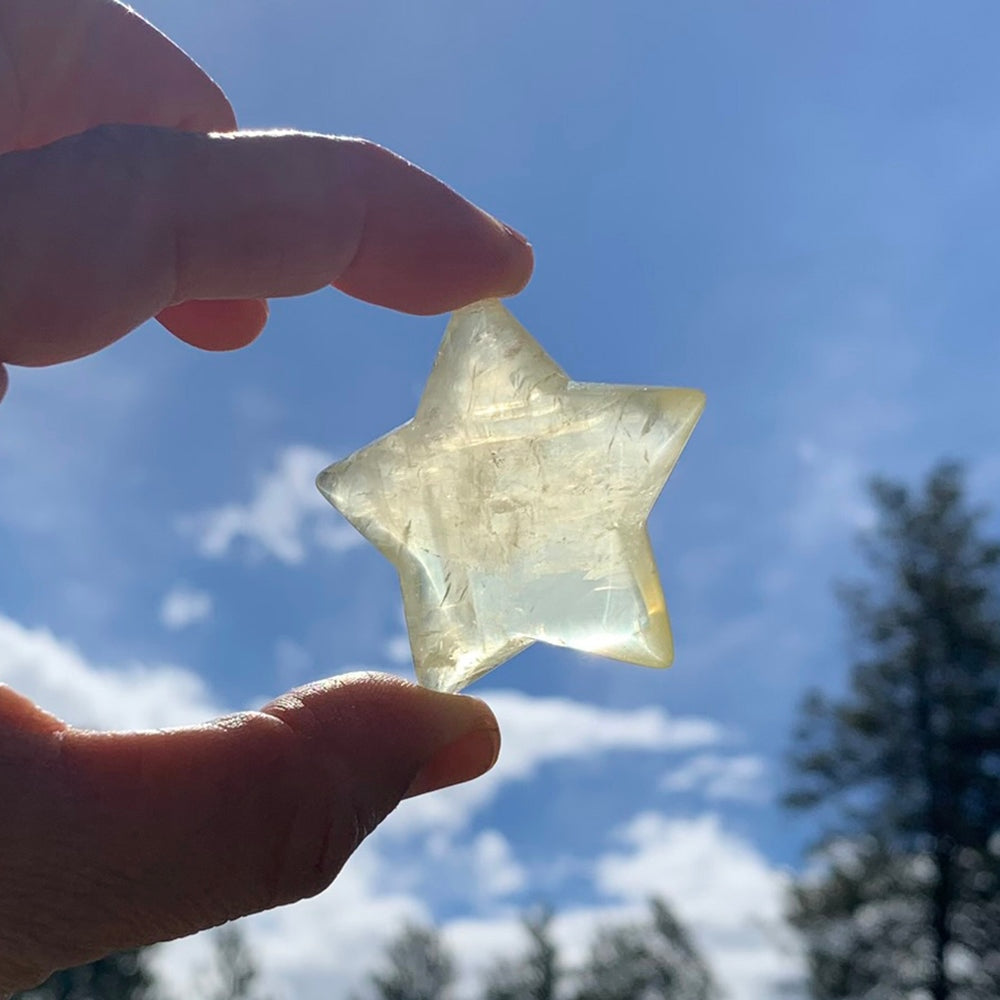 A pale golden citrine star held between fingertips against a blue sky, glowing with light shining through it