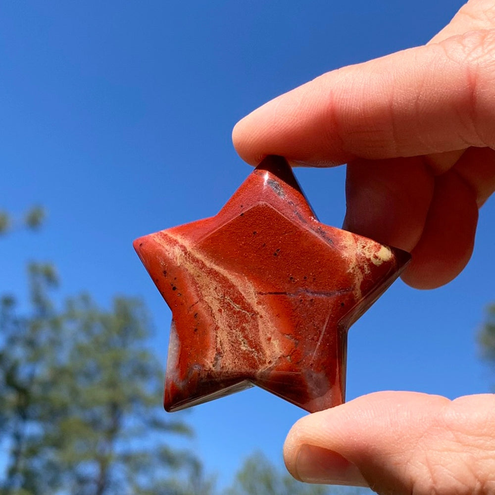 The red jasper star held between thumb and finger against a blue sky, its cream veining catching the light