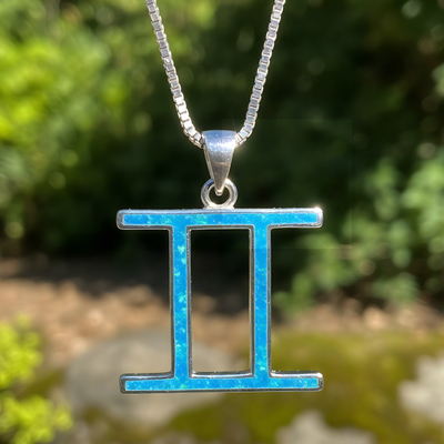 The Gemini glyph pendant inlaid with blue opal, hanging from a sterling silver box chain in a sunlit garden