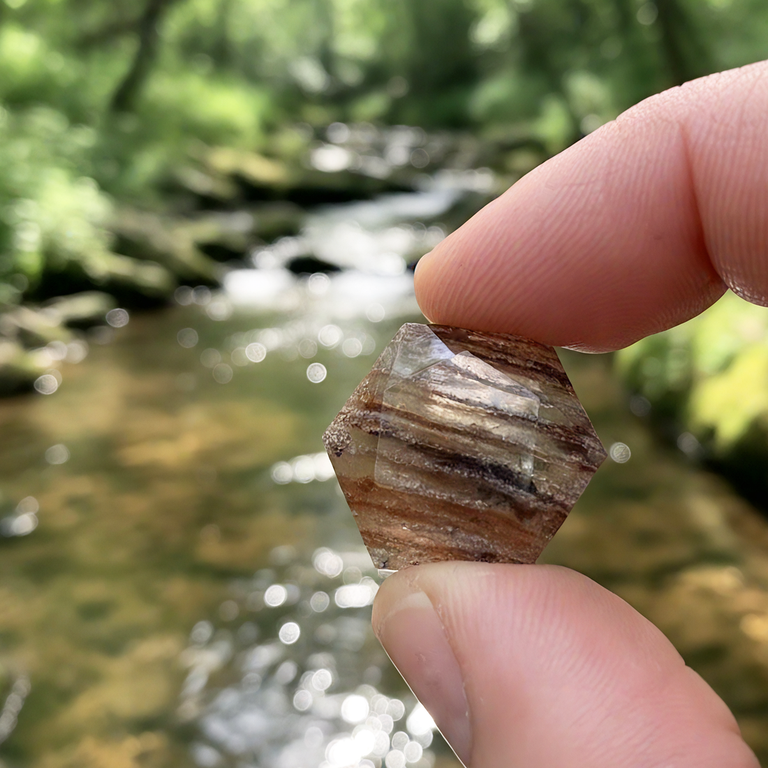The faceted rutilated quartz pinched between finger and thumb above a forest stream, showing its striped copper bands