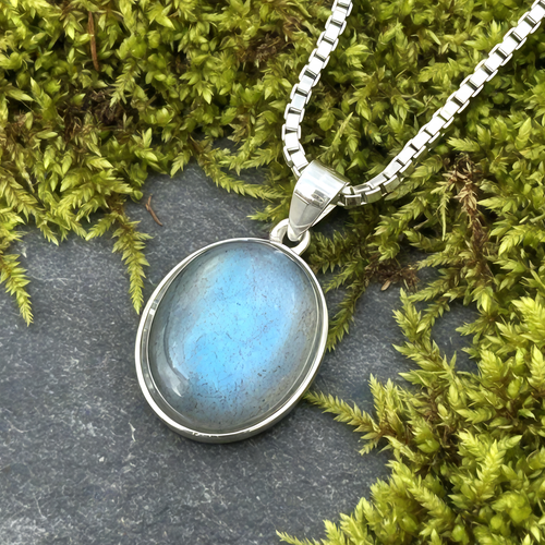 Close-up of the oval rainbow moonstone pendant glowing soft blue in a sterling silver bezel, on a box chain over moss
