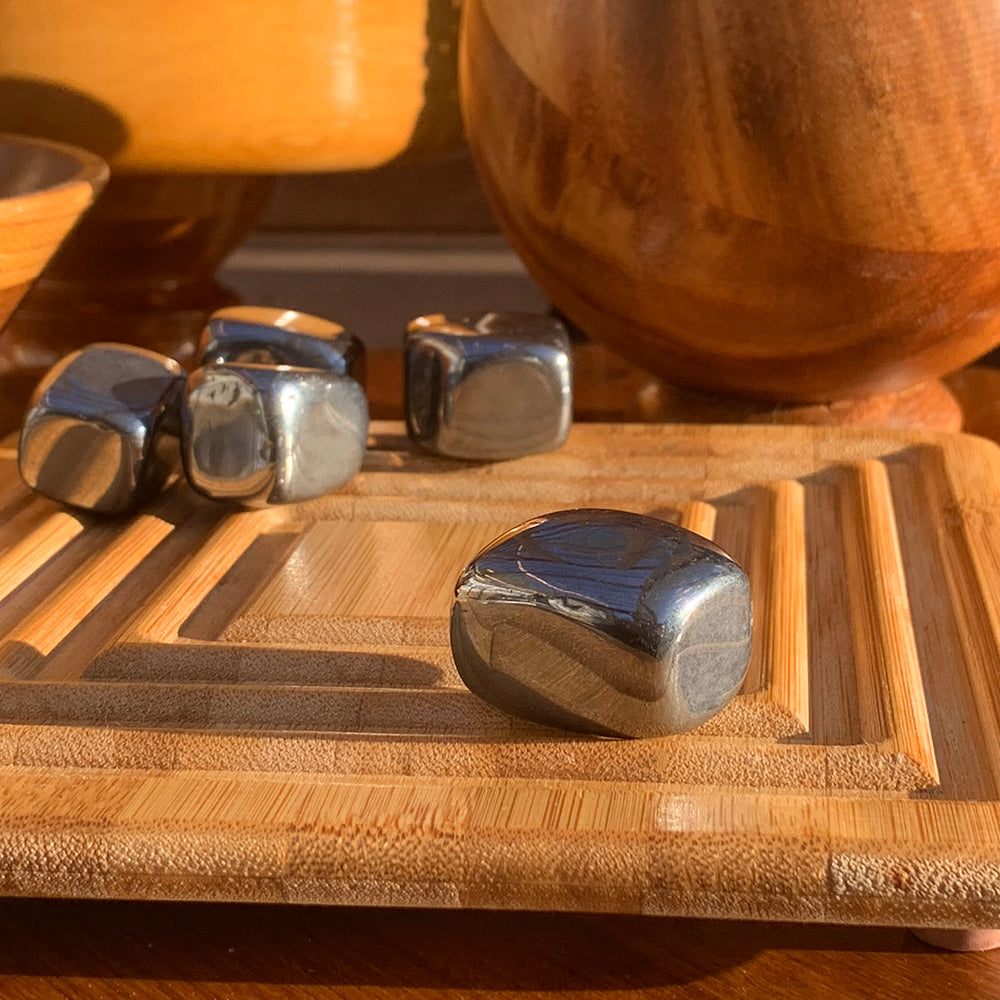 A polished silver hematite cube on a bamboo tray in warm light, with more cubes behind it
