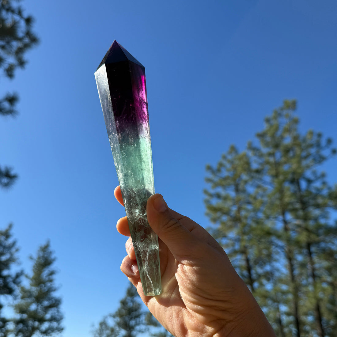 A hand holding the faceted fluorite generator up to a blue sky, deep purple at the point fading into sea-green