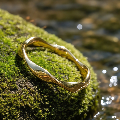 Gold Wave Ring resting on green moss beside a sparkling stream, its band rising and falling in a gentle wave