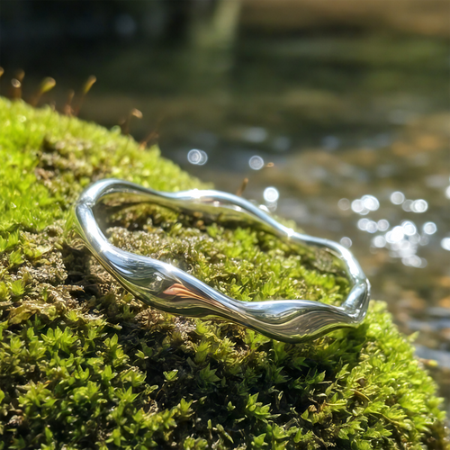 Silver Wave Ring resting on green moss beside a sunlit stream, its band rising and falling in a gentle wave