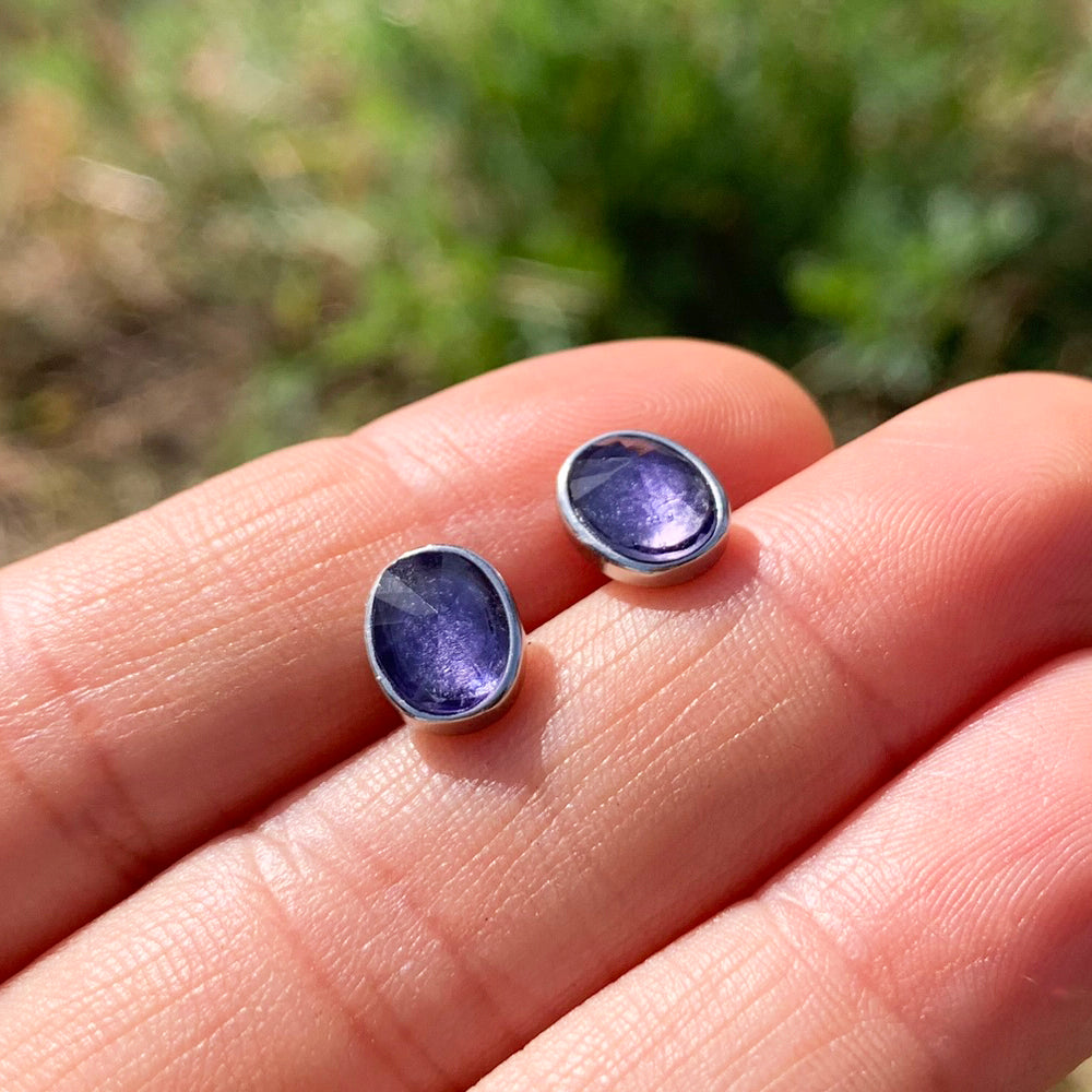 Two oval faceted amethyst studs in sterling silver bezels resting on fingertips in sunlight