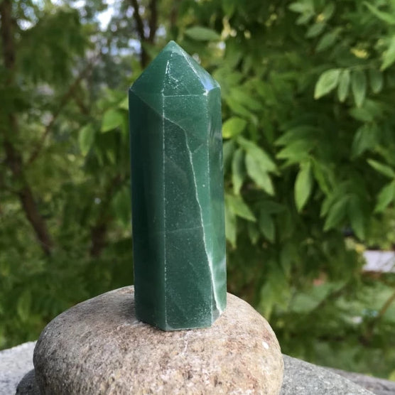 A polished green aventurine point standing on a rounded stone outdoors