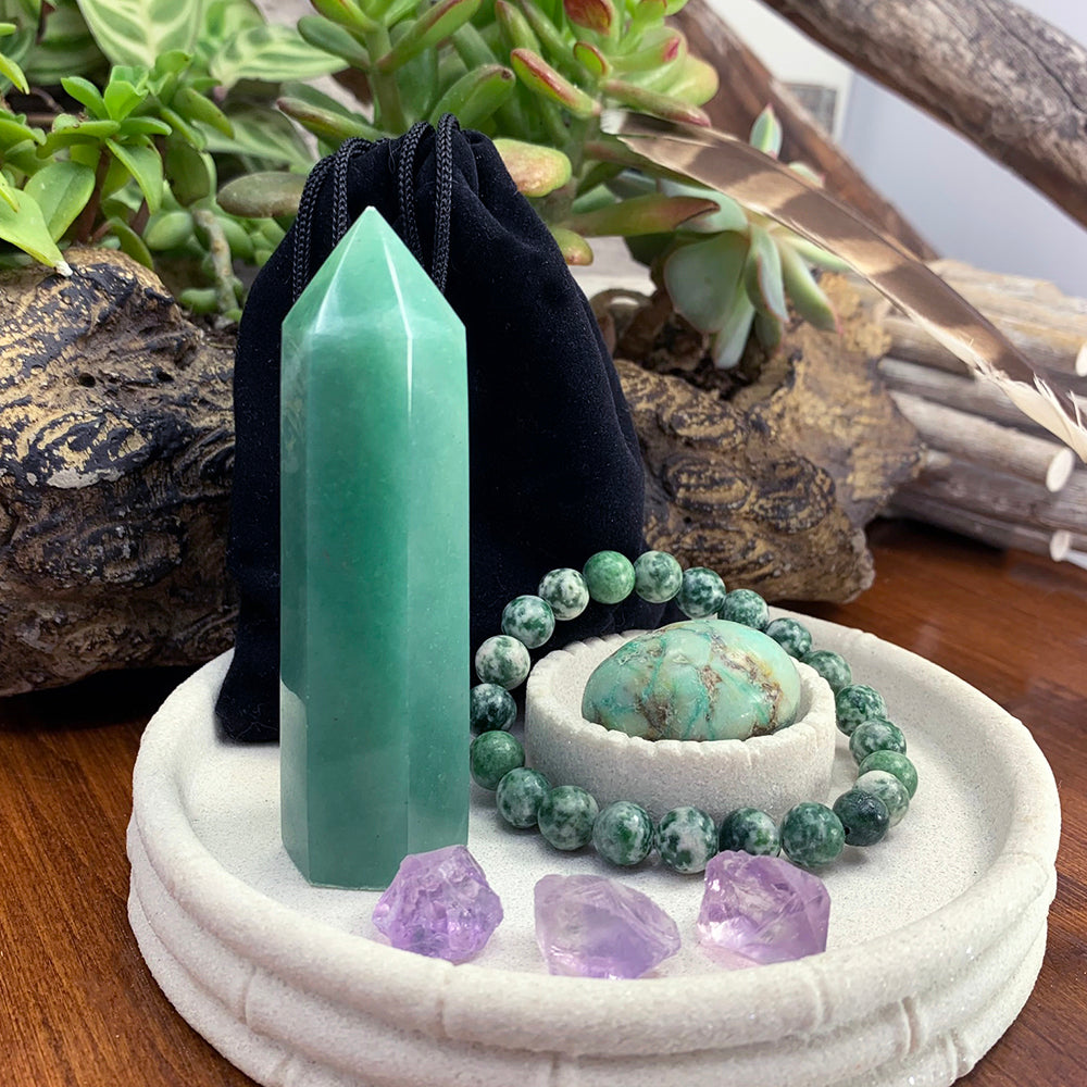 A green aventurine point, a green spot jasper bead bracelet around a phoenix jasper ellipse stone, three rough amethyst pieces and a black velvet pouch in a white dish