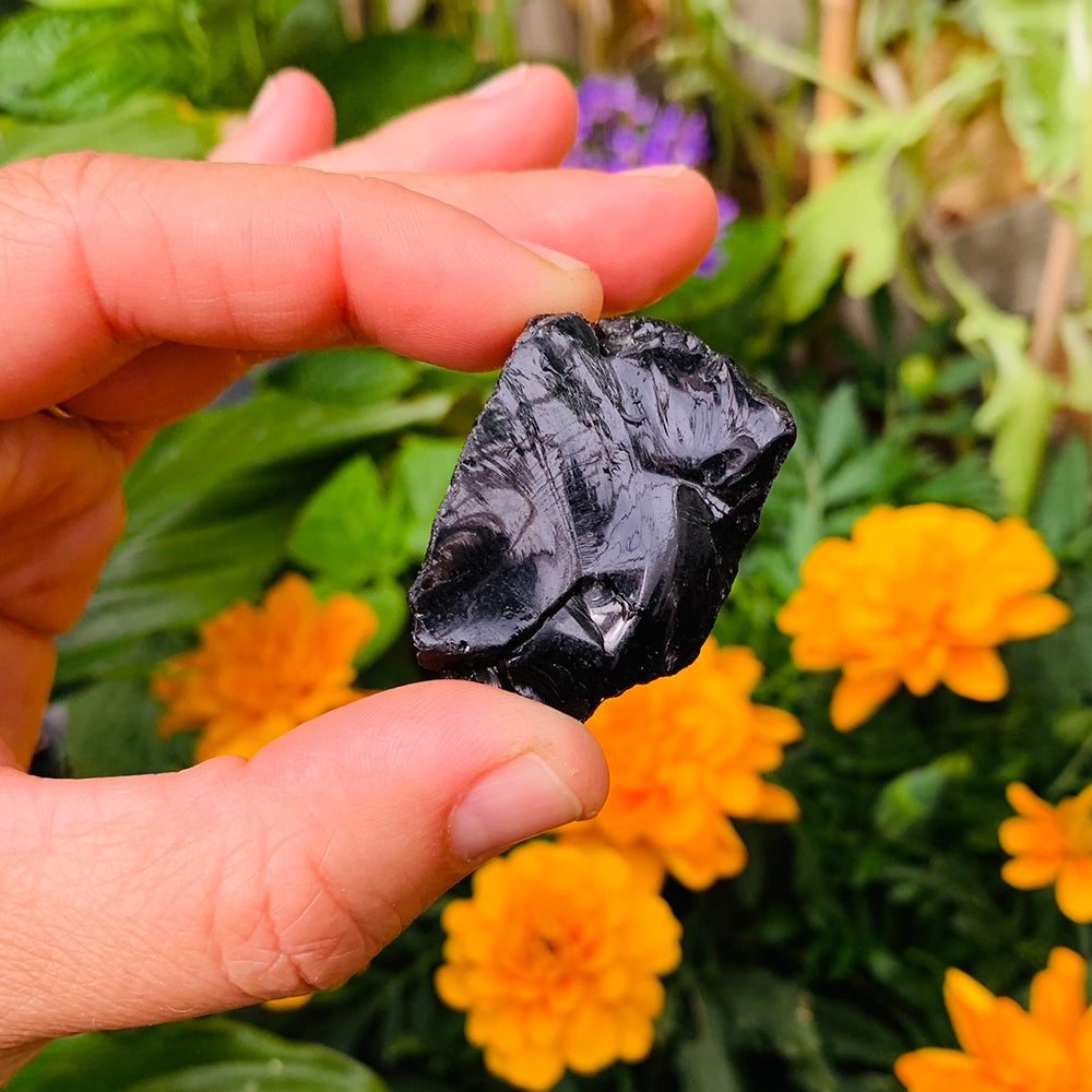 A glossy raw black obsidian stone held between thumb and fingertip in front of orange marigolds