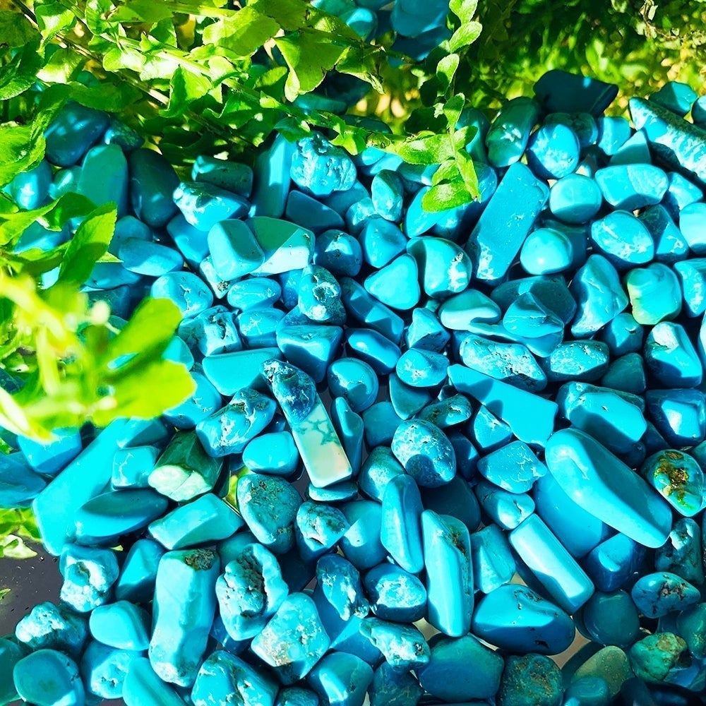 A sunlit heap of bright blue howlite mini gemstones with fine pale veining, beside green leaves