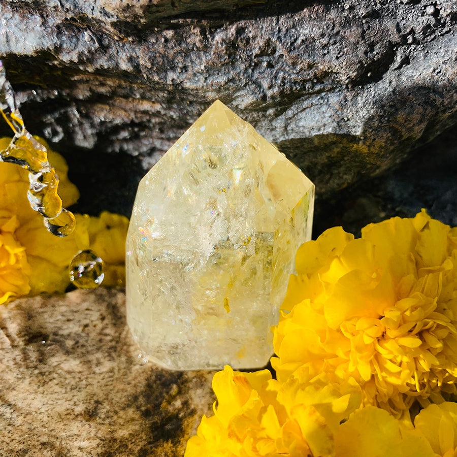 A short, thick pale-gold citrine generator glowing in sunlight on a stone ledge beside yellow marigolds