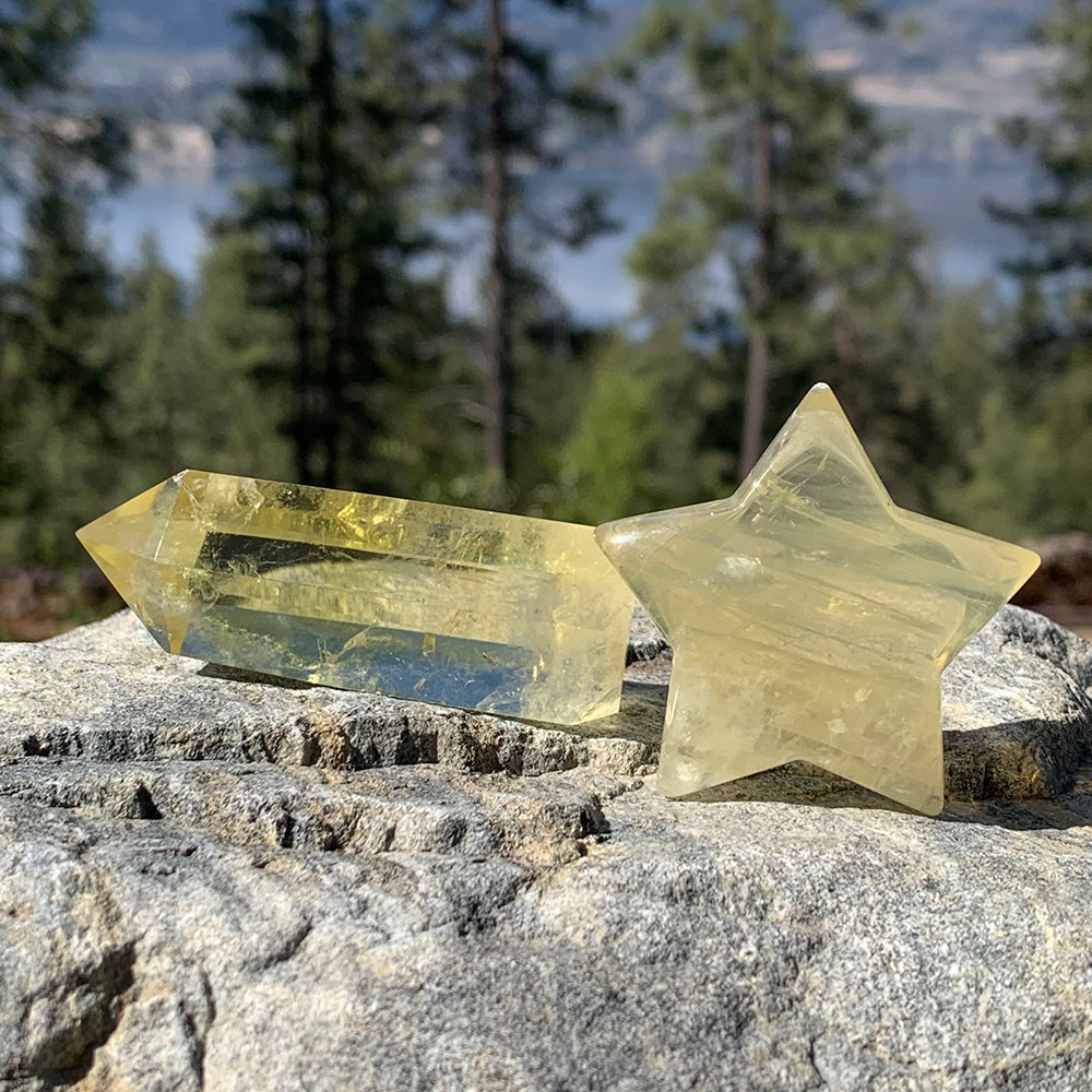 A pale golden citrine point lying beside a polished citrine star on a granite rock, with pine trees and a lake behind