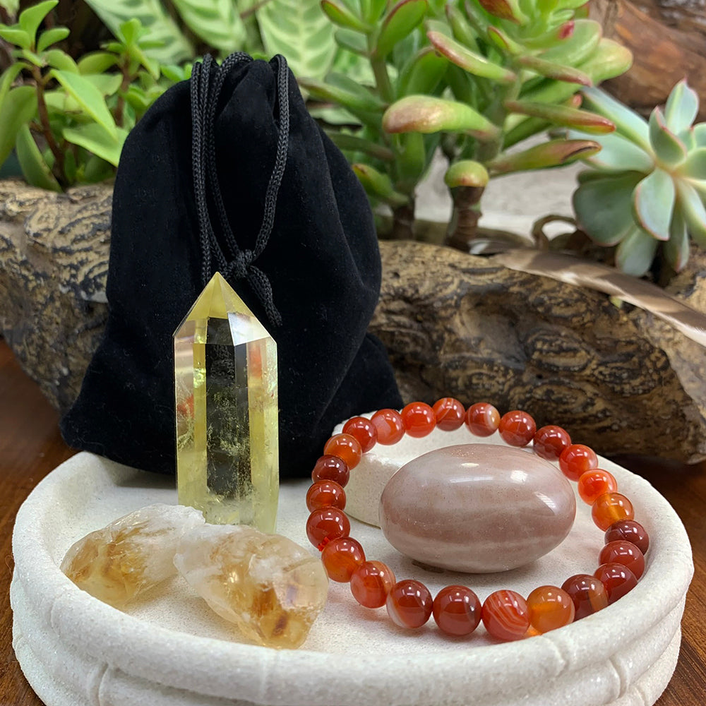 A citrine point, two rough citrine shards, a stripe red agate bead bracelet around a polished ellipse stone and a black velvet pouch in a white dish