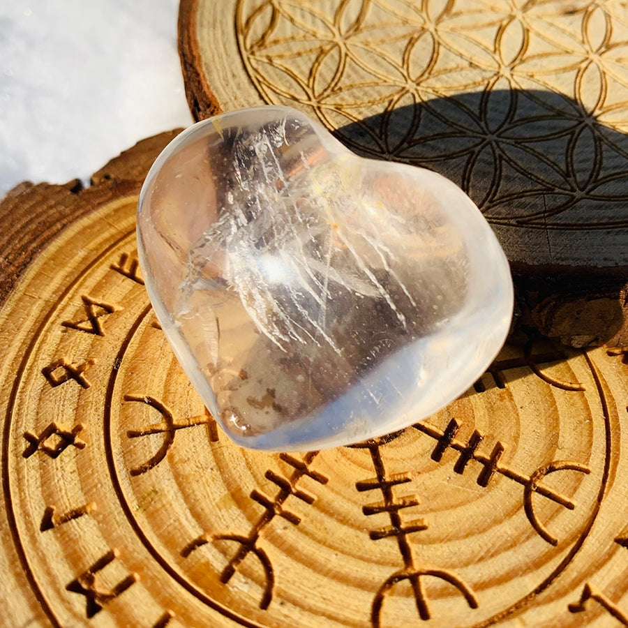 A polished clear quartz heart with milky veils and inclusions, resting on a carved wooden plate in sunlight
