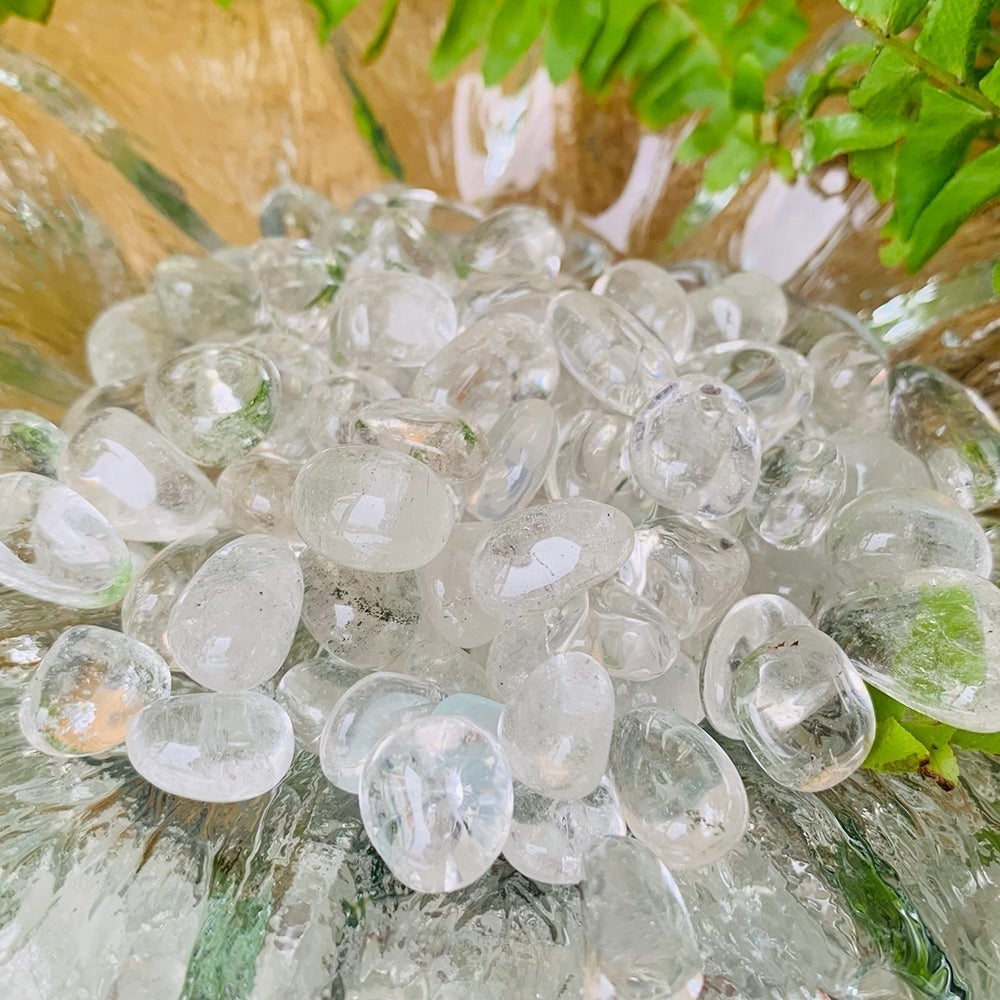 Close-up of a heap of glassy tumbled clear quartz mini gemstones in a glass bowl beside green fern leaves