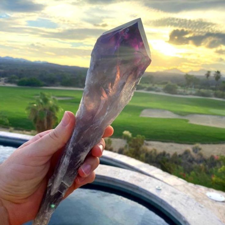 Dark amethyst crystal wand held up against a sunset, light glowing through its violet tip