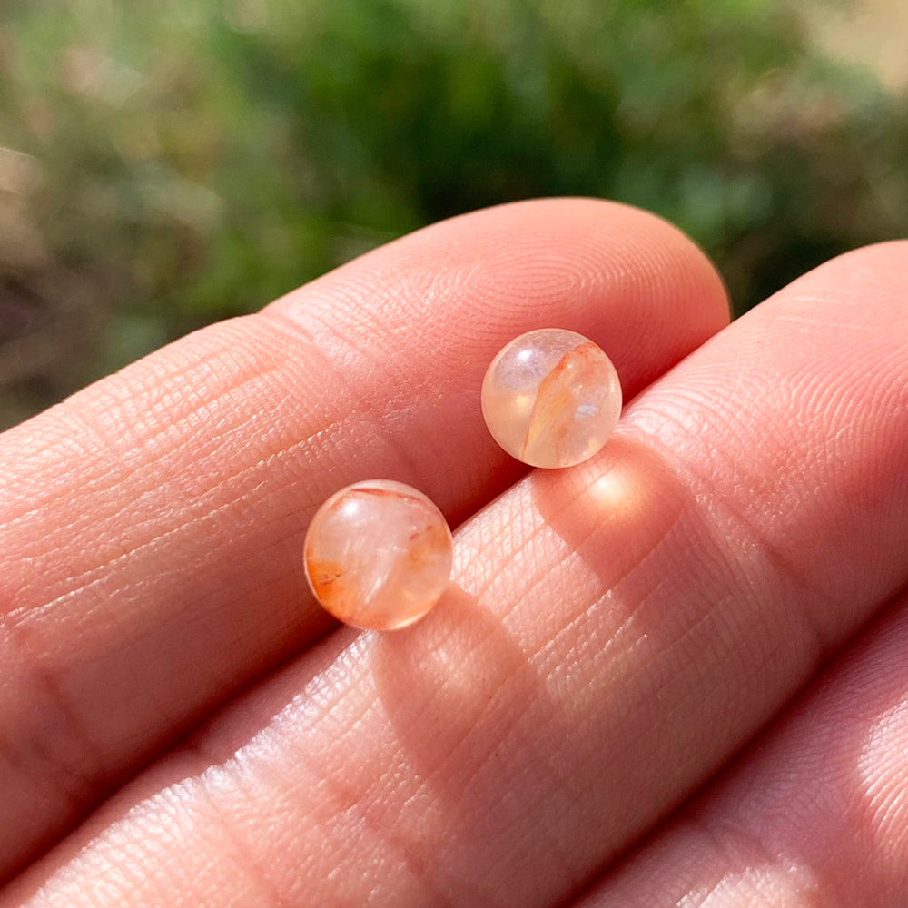 Two round polished fire quartz studs, clear with streaks of red-orange inside, resting on fingertips in sunlight
