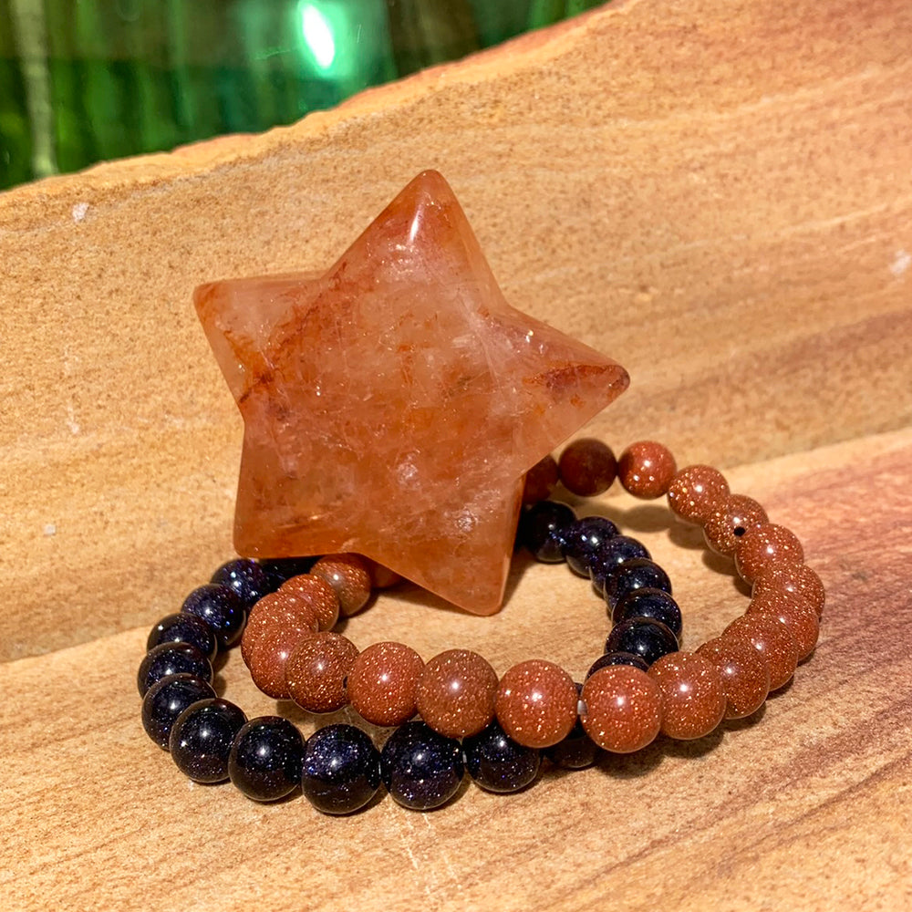 A peach and rust fire quartz star resting on a coppery red sandstone bracelet and a midnight blue sandstone bracelet on sandstone