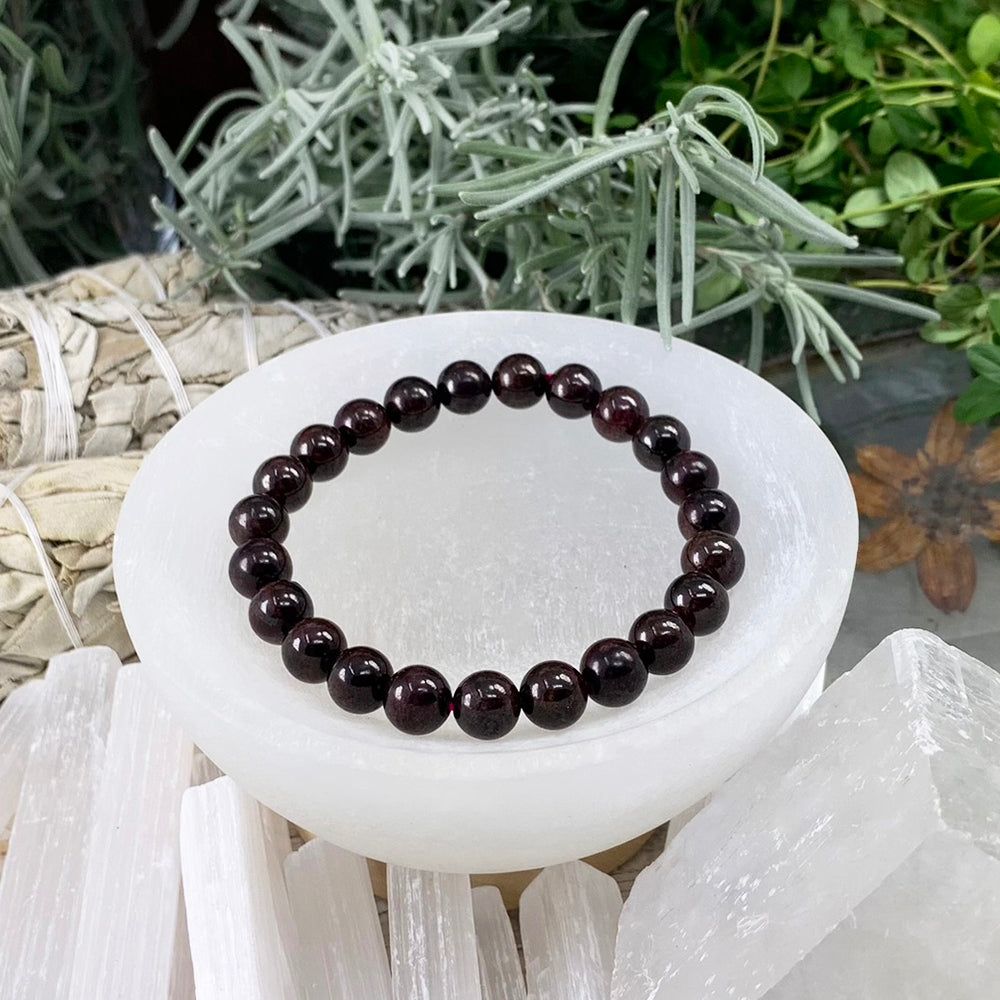 A garnet mala bracelet of round, glossy deep-red beads resting in a white selenite bowl
