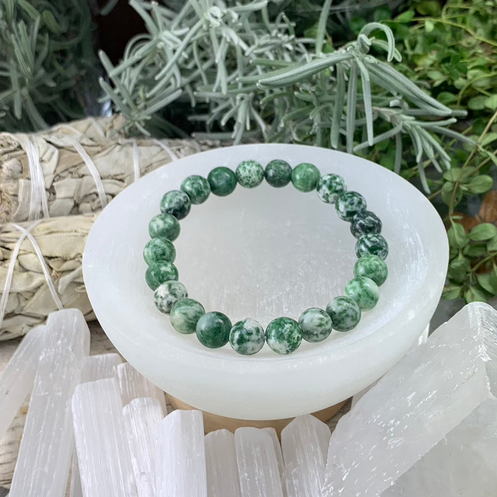 One green spot jasper mala bracelet of round green beads speckled with white, resting in a white selenite bowl