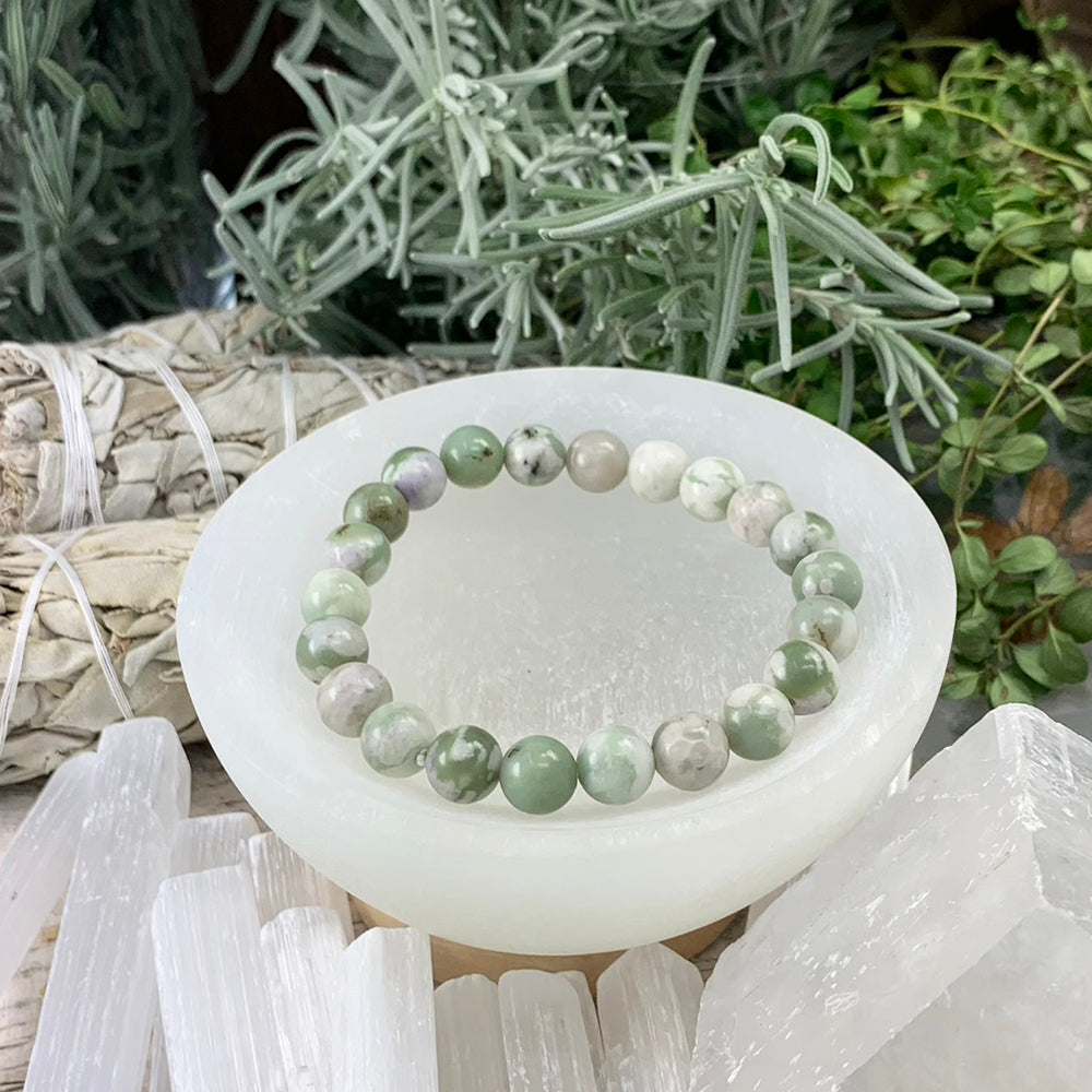 A jade bead bracelet in mottled sage green, cream and soft lavender resting in a selenite bowl