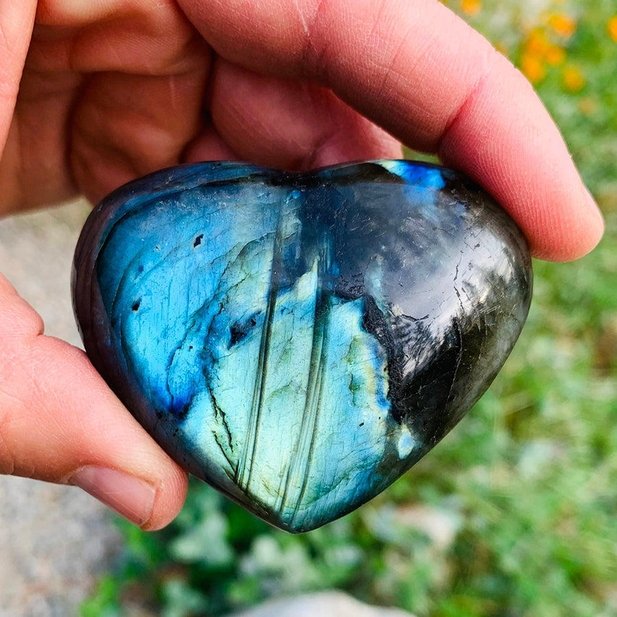A labradorite heart held between finger and thumb, its face flashing blue and teal