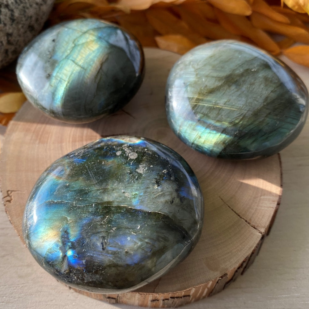 Three labradorite palm stones on a wooden round, each flashing a different mix of blue, green and gold
