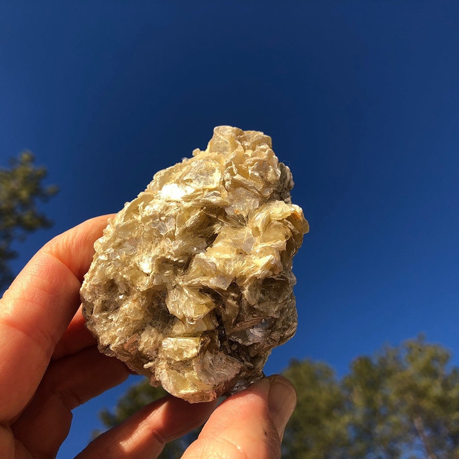 A rough golden mica cluster held up against a blue sky, its stacked flakes catching the sunlight