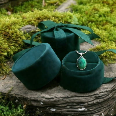 The malachite necklace nestled in an open round green velvet jewelry box on a mossy stone, beside a ribboned velvet box
