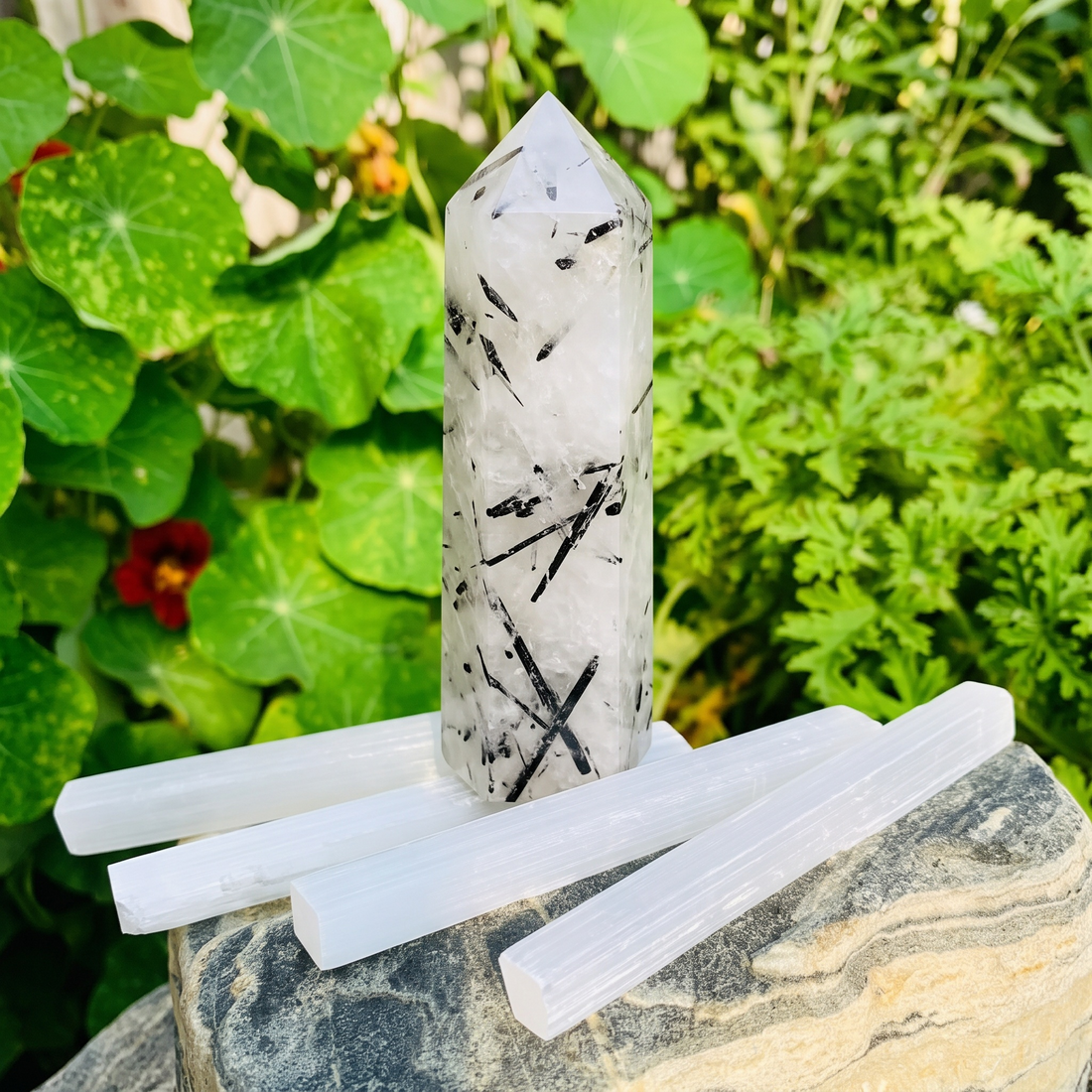A clear quartz point streaked with black tourmaline needles standing on four white selenite sticks on a garden rock