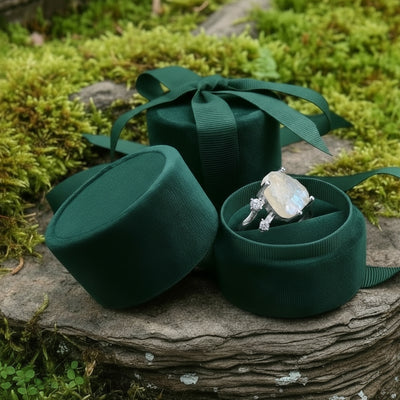 The moonstone ring nestled in an open round velvet ring box beside its lid on a mossy stone