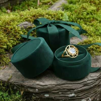 The solar glow ring nestled in an open round green velvet ring box beside its lid and ribbon on a mossy stone