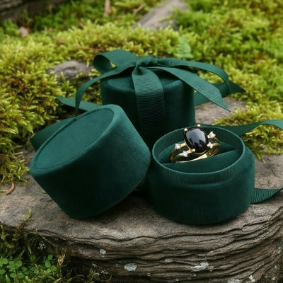 The black onyx and gold ring nestled in an open round green velvet jewelry box on a mossy stone