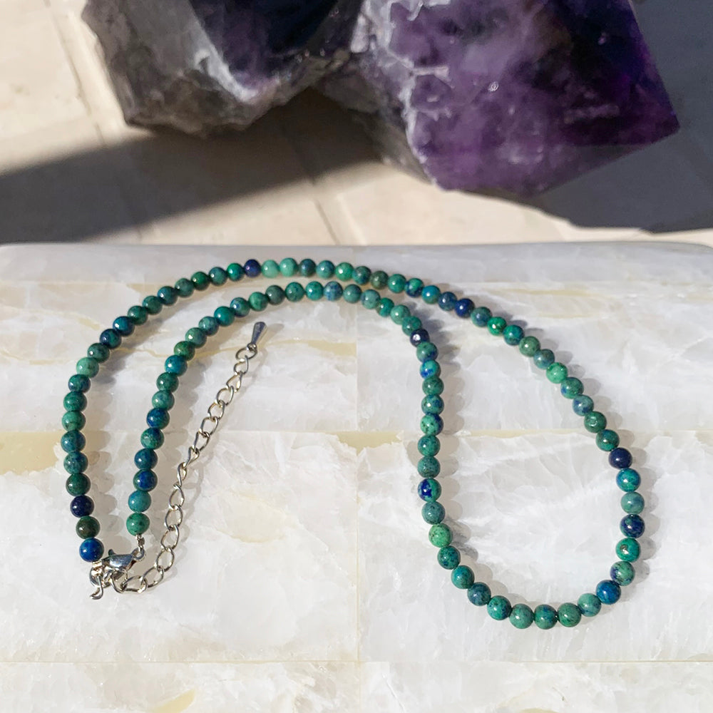 A phoenix lapis bead necklace in mixed green and deep blue beads, laid out with its silver clasp and extender chain on white stone