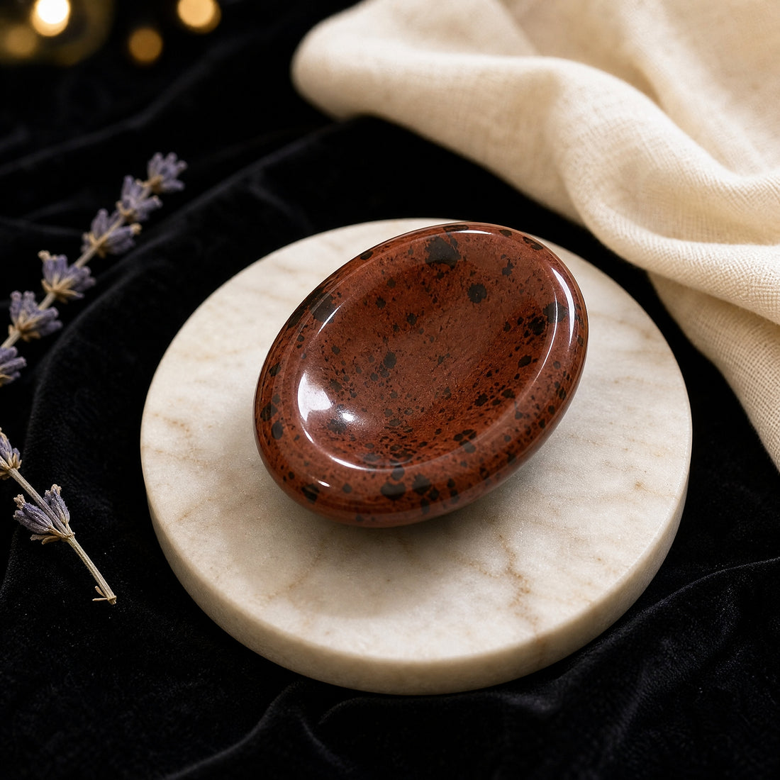 Polished oval red obsidian worry stone, deep red-brown with black flecks and a shallow thumb groove, on a marble dish