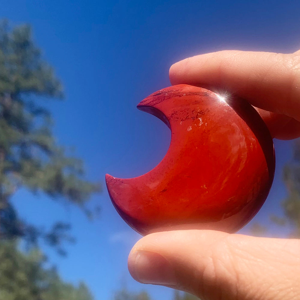 A polished red jasper crescent moon held between thumb and fingers against a blue sky