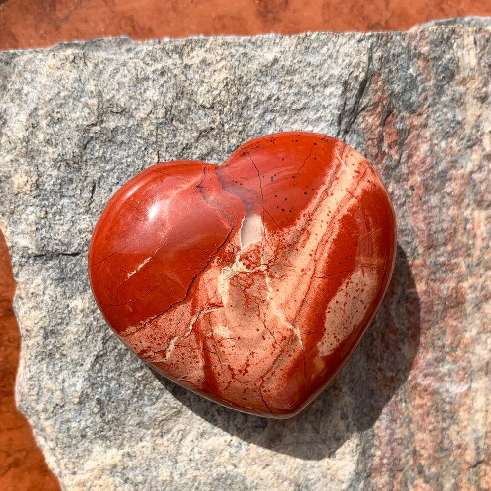 A polished red jasper heart swirled with brick red and cream, resting on sunlit stone