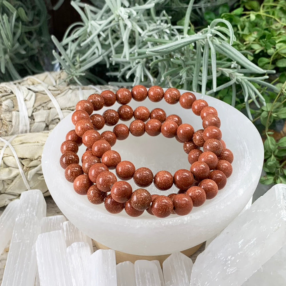 Red sandstone mala bracelets of glossy copper-red beads flecked with sparkles, stacked in a white selenite bowl