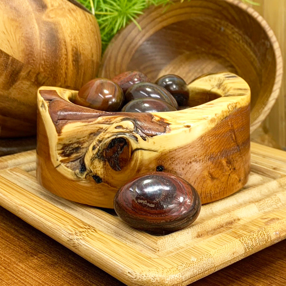 A polished red tiger’s eye ellipse with deep red and brown bands resting in front of a carved wooden bowl of more red tiger’s eye stones