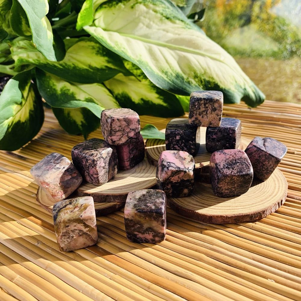 Dusty pink rhodonite tumbled cubes veined with black, stacked on wood slices on a bamboo mat beside a leafy plant