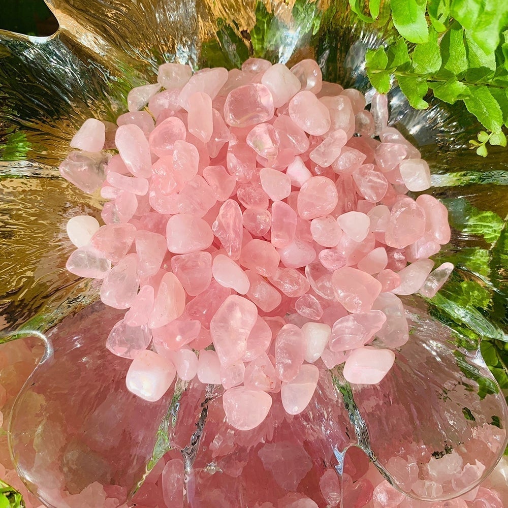 A clear glass flower dish heaped with pink tumbled rose quartz mini gemstones, framed by green leaves