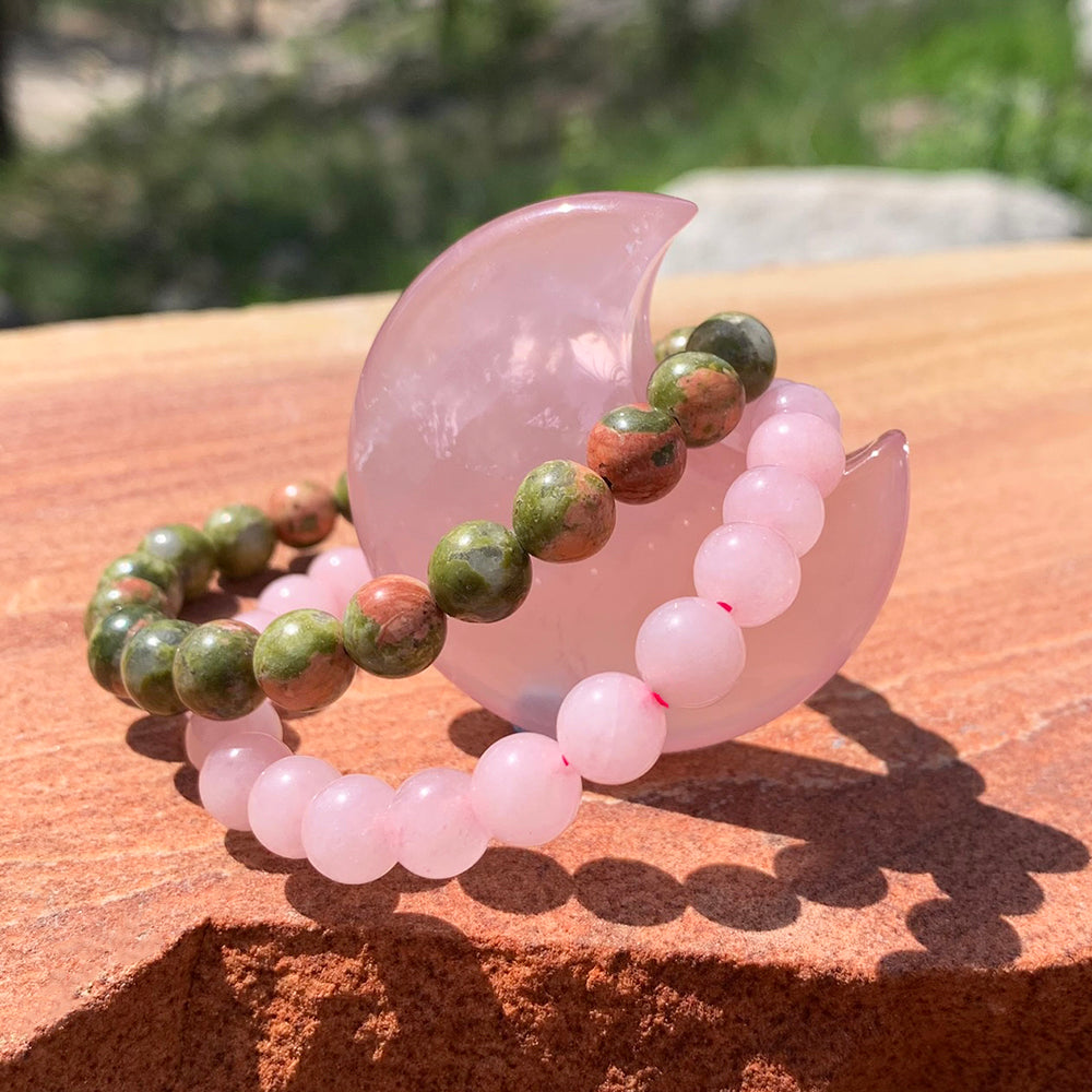 A pink rose quartz mala bracelet and a green and pink unakite mala bracelet draped over the standing rose quartz crescent moon in the sun