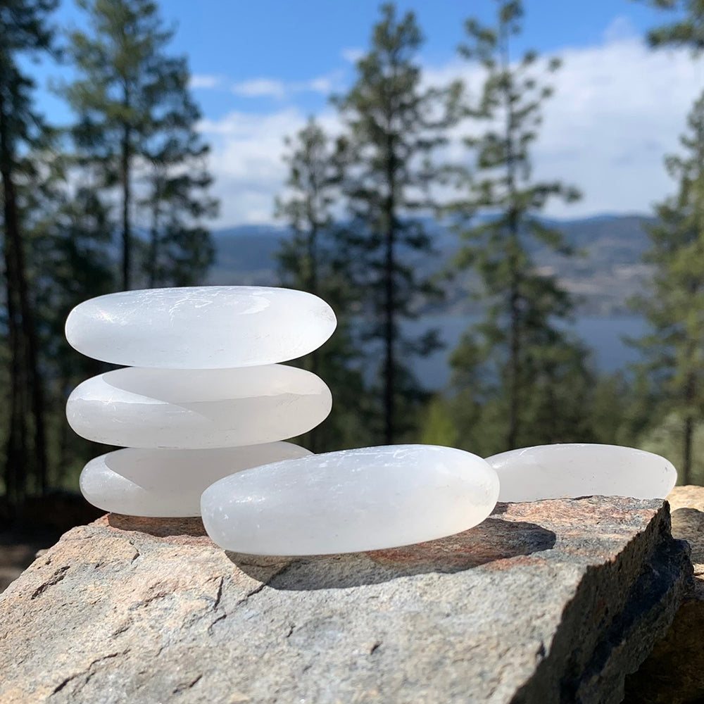 Five white selenite palmstones stacked and laid on a sunlit rock above a lake and pine trees