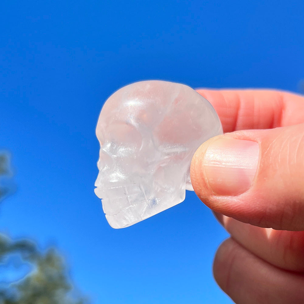 A small clear quartz skull held between fingertips against a bright blue sky, light passing through it