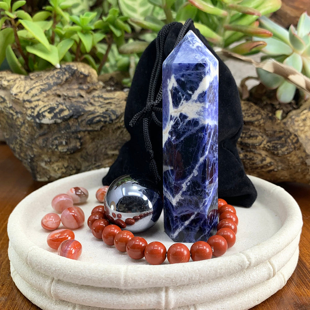 A blue and white sodalite point, a silver hematite ellipse stone, a red jasper bead bracelet and red carnelian minis in a white dish with a black velvet pouch