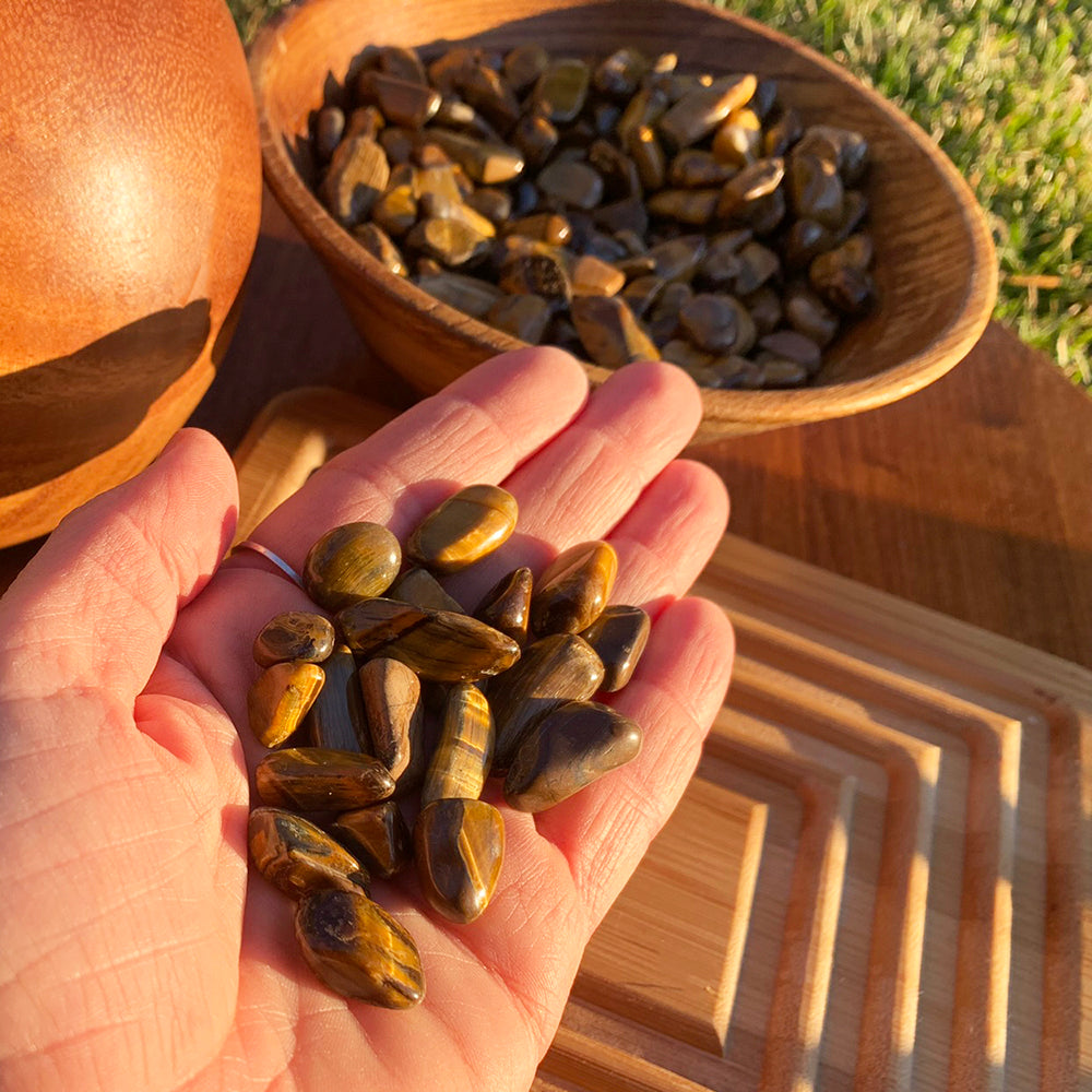 A small handful of tiger’s eye mini gemstones in an open palm, with a wooden bowl of more stones behind