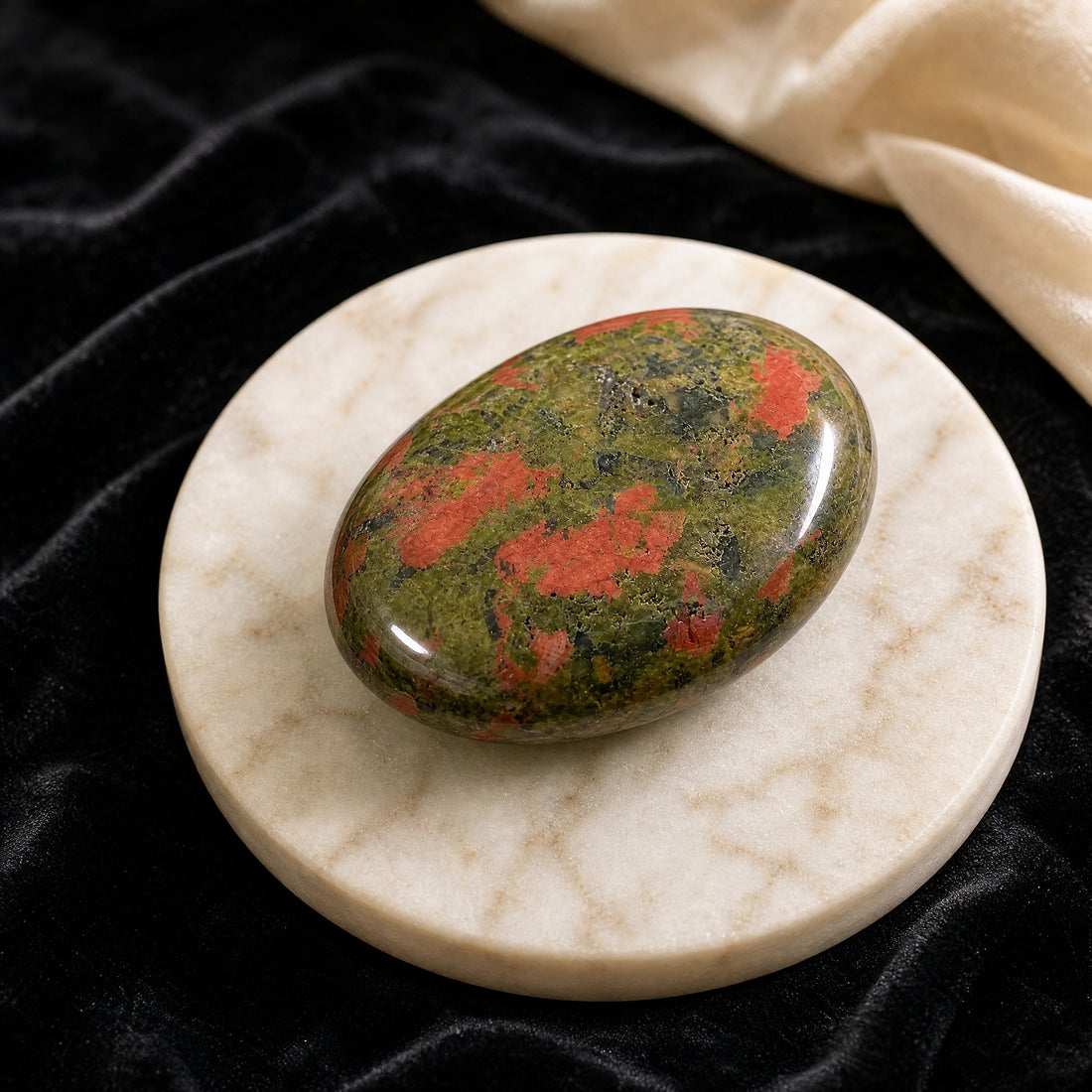 A polished oval unakite palmstone, olive green flecked with salmon pink, on a pale marble dish