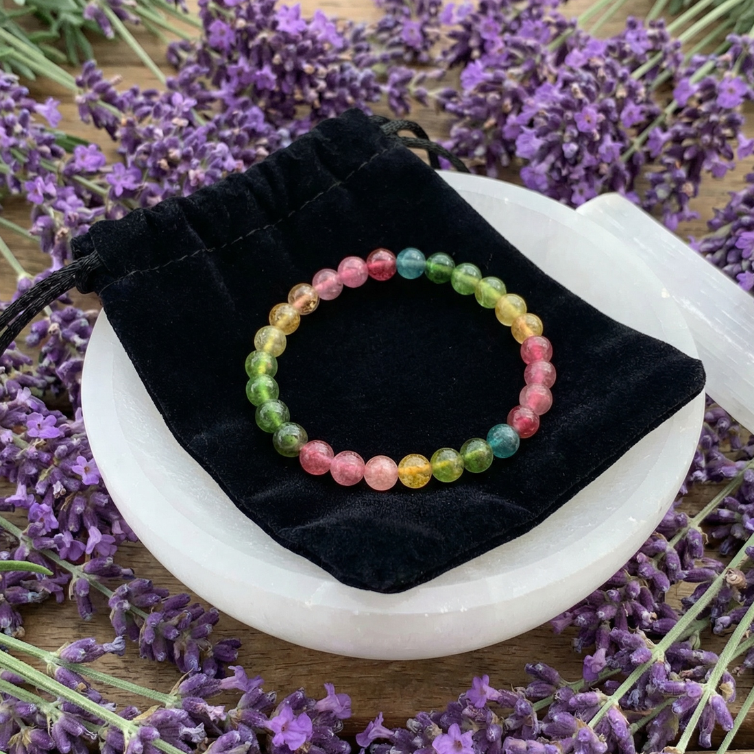 The watermelon tourmaline bracelet laid on its black velvet drawstring pouch, on a white selenite plate among lavender