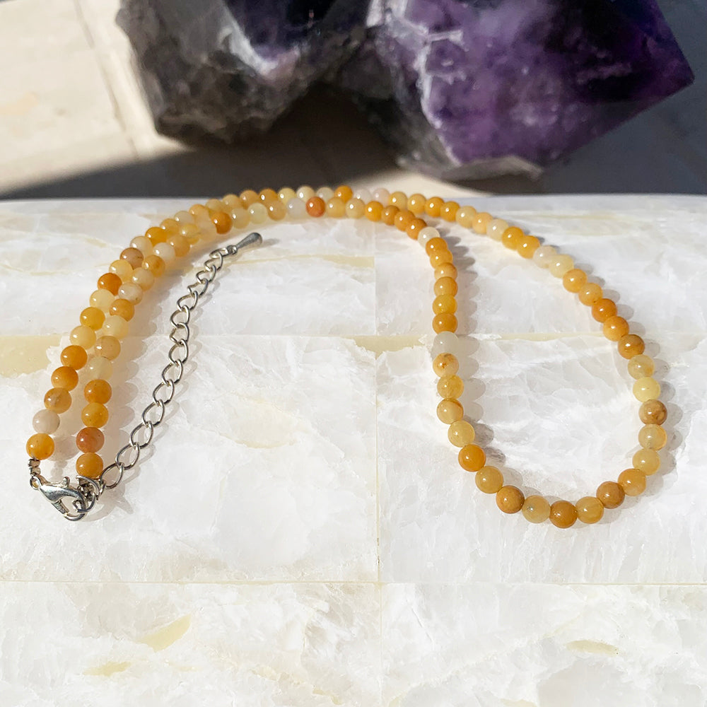 A yellow aventurine bead necklace in honey and golden tones with its silver clasp and extender chain on white stone, an amethyst cluster behind